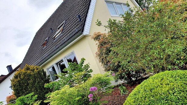 Une maison avec un toit en pente, une clôture blanche et divers arbustes et fleurs.