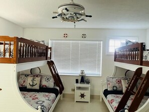 A room with two bunk beds, a small white nightstand with a ship wheel decoration, and a ceiling fan.