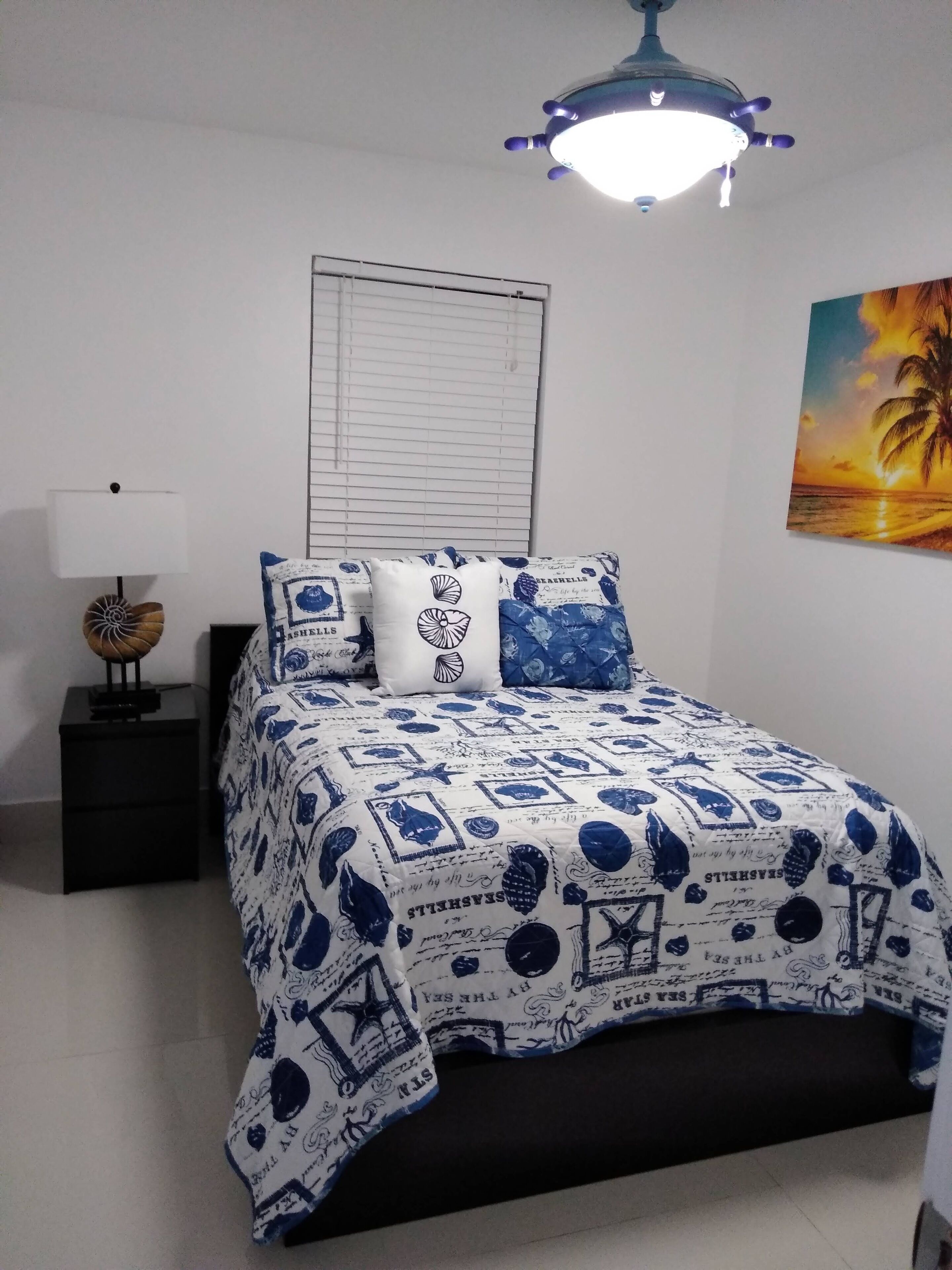 A bedroom with a bed, a nightstand, a lamp, a window with blinds, and a painting of a sunset.
