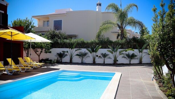A swimming pool with yellow lounge chairs and a white umbrella, surrounded by a wooden deck and greenery. - Sunshine Villa (Faro)