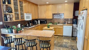 A kitchen with wooden cabinets, a granite countertop, a microwave, a stove, and a refrigerator. There are three bar stools and a small island with a plant. - Cheerful Single Story 3-bedroom home w pool table (Santa Clarita)