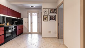 A modern kitchen with red cabinets, a black countertop, and a tiled floor. There are framed artworks on the wall and a door leading to another room. - Aurora Villa II with private Jacuzzi - Naxos (Agios Prokopios)