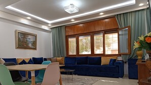 A modern living room with a blue sofa, a dining table with chairs, and a large window with curtains. - Luxury Family House with Garden View Casablanca (Dar-el-Beida)