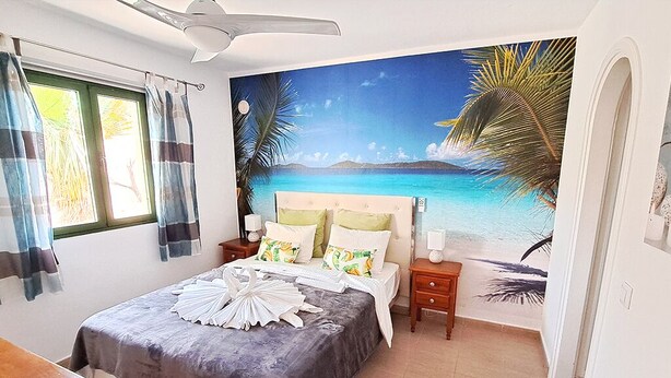 A bedroom with a bed, a nightstand, a wooden table, a ceiling fan, and a beach mural on the wall. - Casa Helena (Costa Calma)