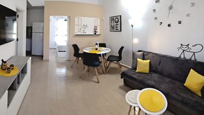 A modern living room with a sofa, two yellow cushions, a round table with chairs, a flat-screen TV, and a wall clock. - Casa Helena (Costa Calma)