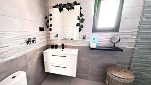 A modern bathroom with a white sink, mirror, and toilet. There is a wicker basket and a small shelf with toiletries. - Casa Helena (Costa Calma)