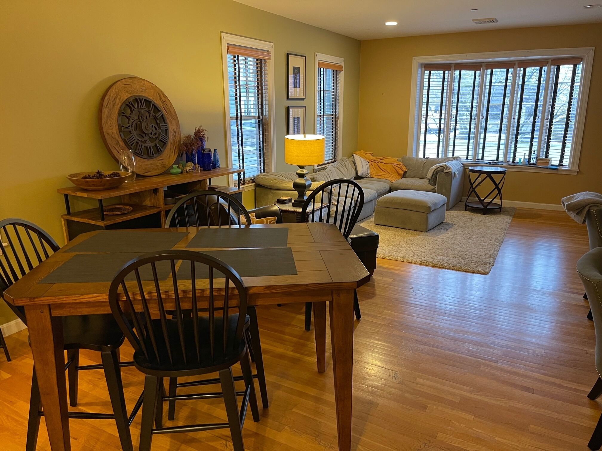 A dining area with a wooden table and chairs, a sofa set, and a large window with blinds.
