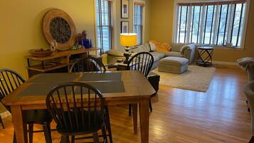A dining area with a wooden table and chairs, a sofa set, and a large window with blinds.
