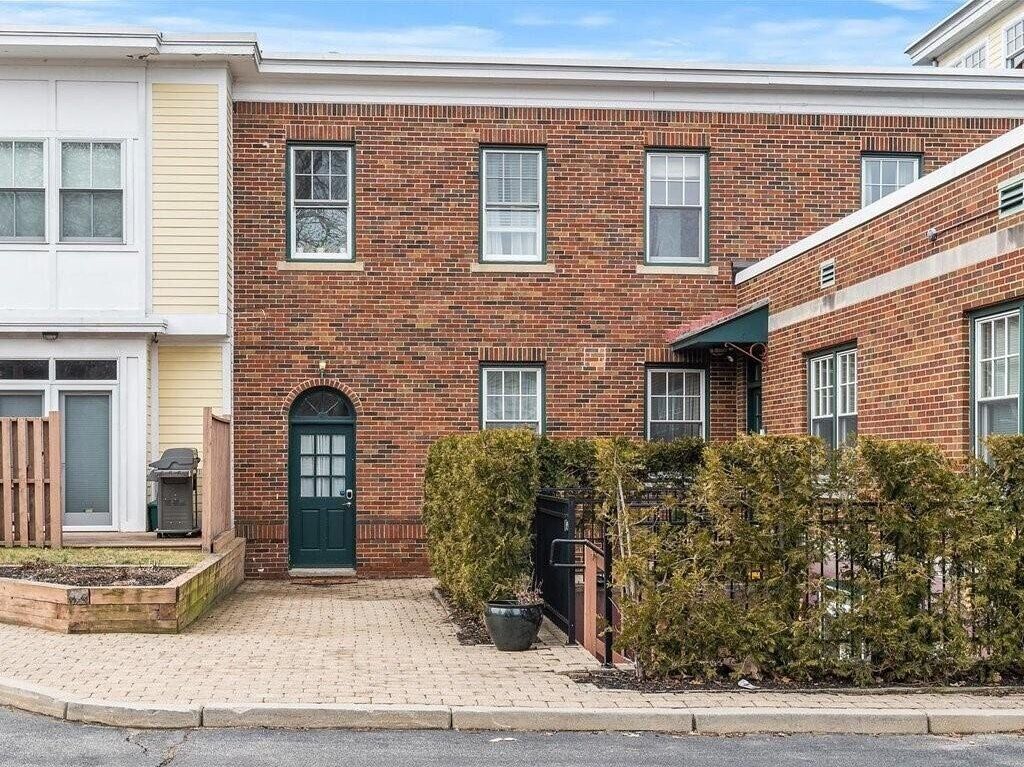 A two-story brick building with a green door, multiple windows, and a small garden area with shrubs.