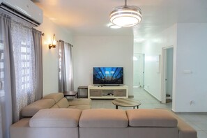 Un salon moderne avec un canapé beige, un téléviseur à écran plat et un luminaire suspendu au plafond.