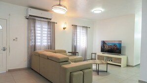 Una sala de estar con un sofá color beige, un televisor sobre un mueble bajo y un ventilador de techo.