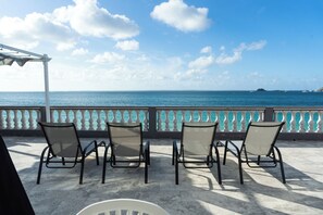 A terrace with four chairs facing a sea view, a white railing, and a clear sky. - Novels Villa is a family-friendly home with the Caribbean Sea as your backyard. (Grand-Case)