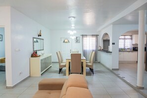 A modern living room with a sofa, dining table, and chairs. There is a kitchen area with cabinets and a countertop visible.
