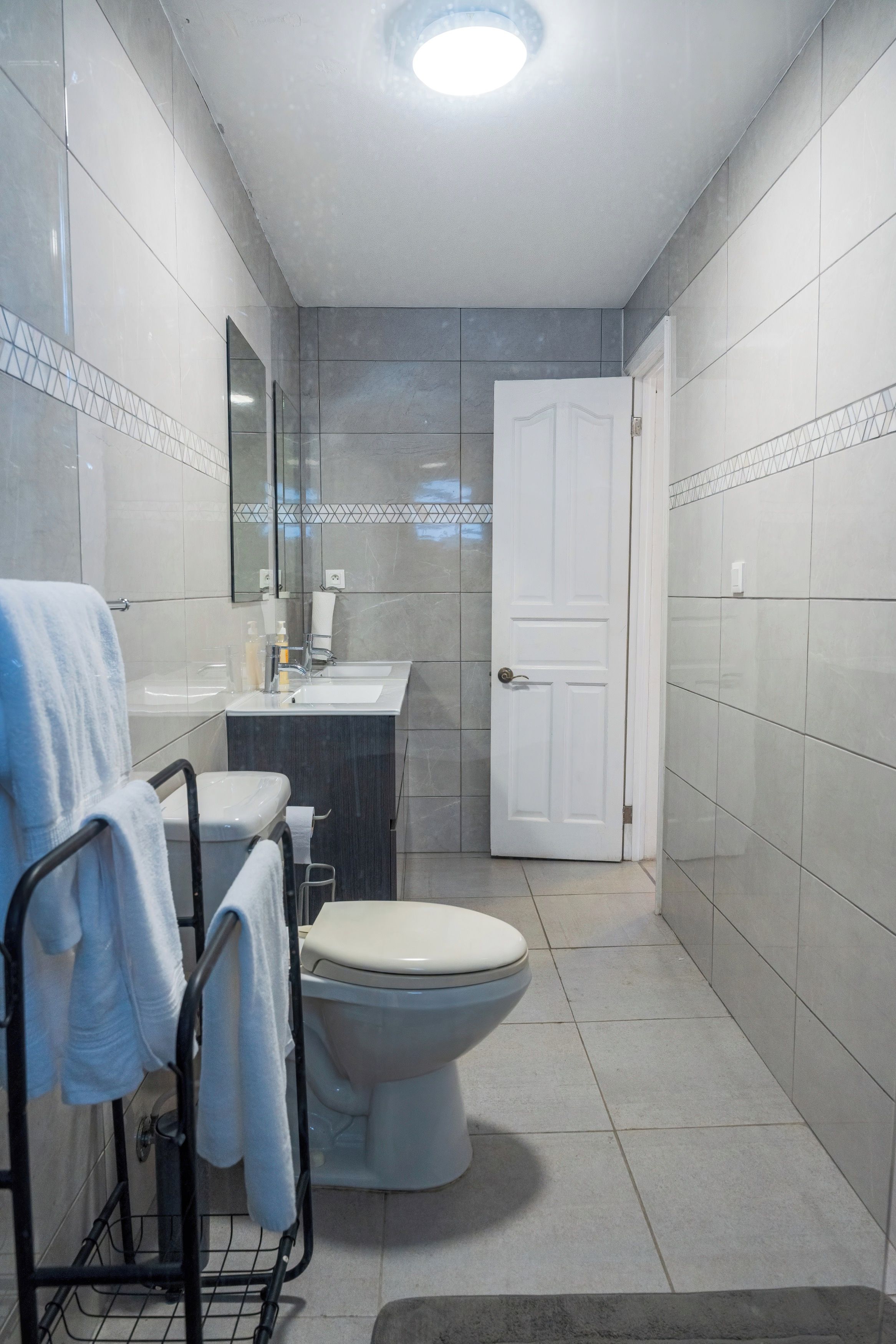 Ein Badezimmer mit Toilette, Waschbecken und Spiegel. Ein Handtuchhalter mit Handtüchern ist vorhanden. Die Wände sind mit Fliesen versehen und es gibt eine Tür.
