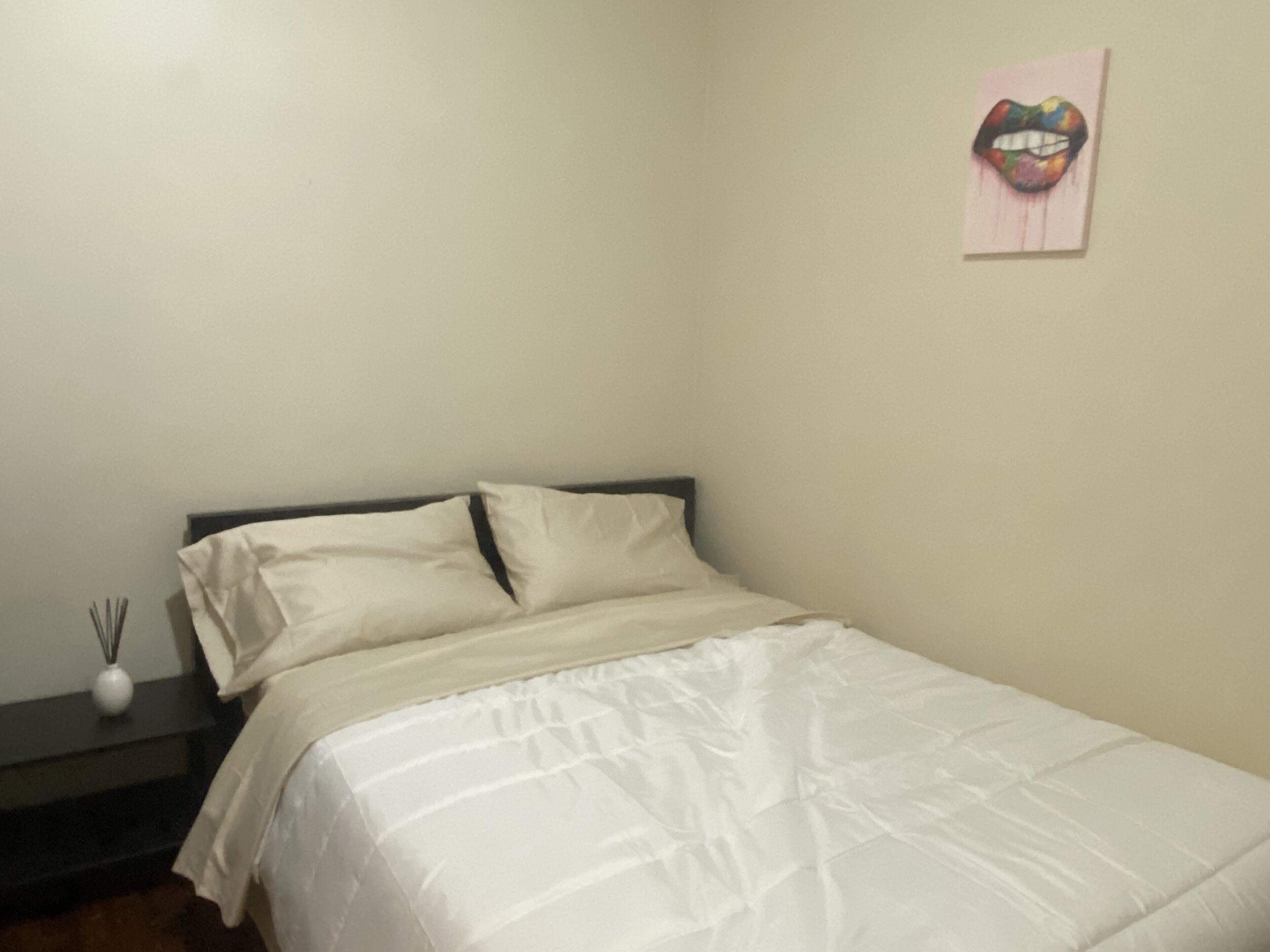 A neatly made bed with white bedding in a simple room with a plain wall and a small artwork of lips.