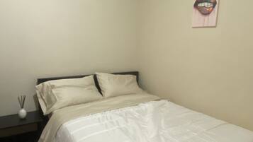 A neatly made bed with white bedding in a simple room with a plain wall and a small artwork of lips.