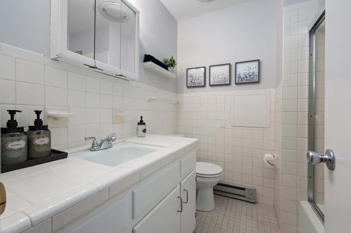 A bathroom with a white sink, toilet, and shower area. There are two framed pictures on the wall, a shelf with toiletries, and a window above the sink.