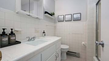 A bathroom with a white sink, toilet, and shower area. There are two framed pictures on the wall, a shelf with toiletries, and a window above the sink.