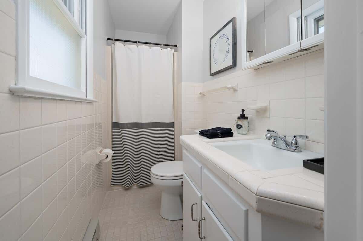 Un bany amb parets de rajola blanca, un vàter, una cortina de bany, un lavabo amb mirall i un suport per a tovalloles.