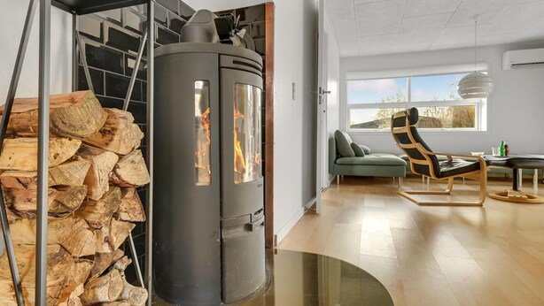 Un salon moderne avec un poêle à bois, une pile de bois de chauffage, un canapé vert et un fauteuil original.