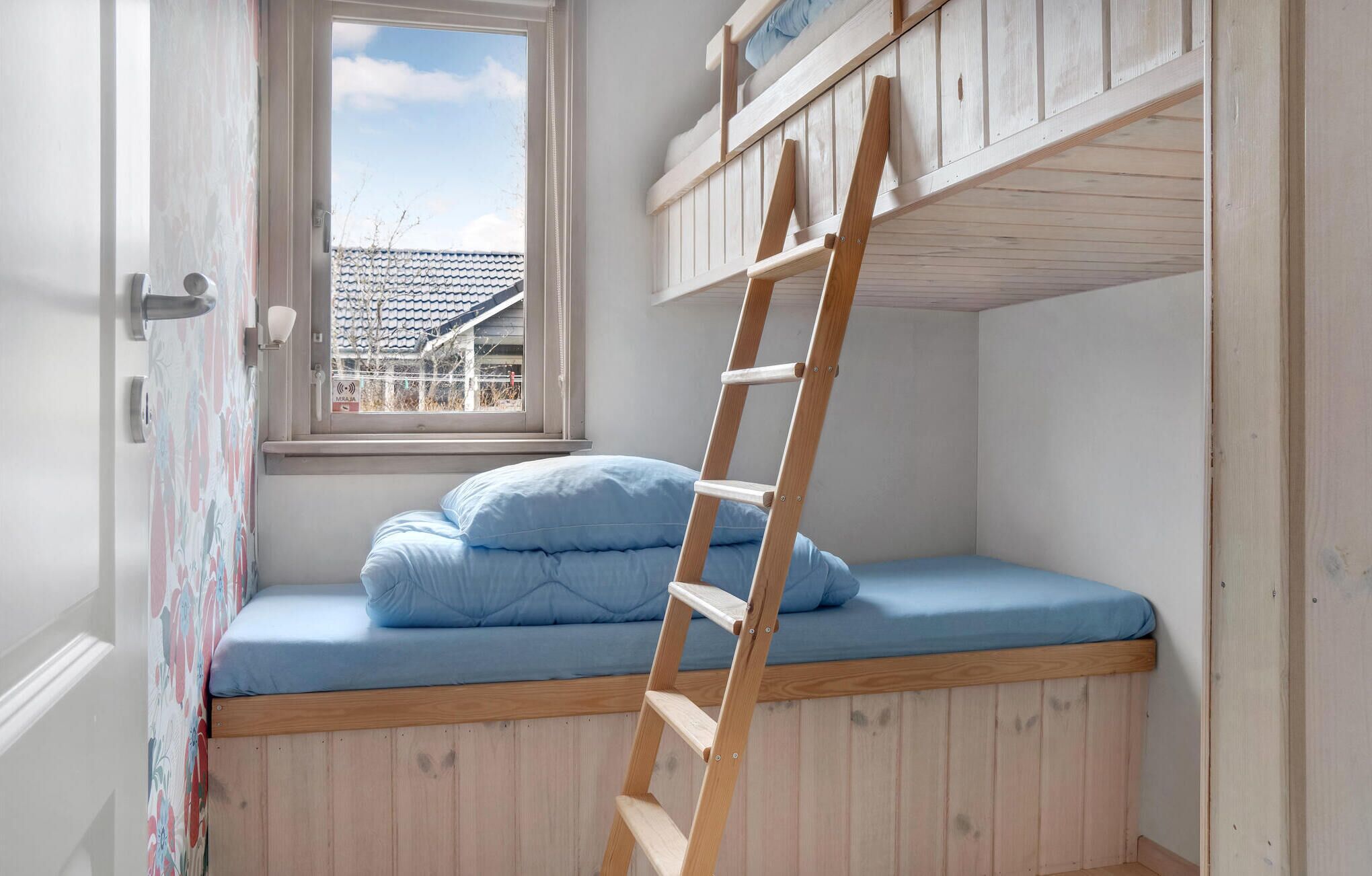 A bunk bed with a wooden ladder, a window with a view of buildings, and a door with a floral pattern.