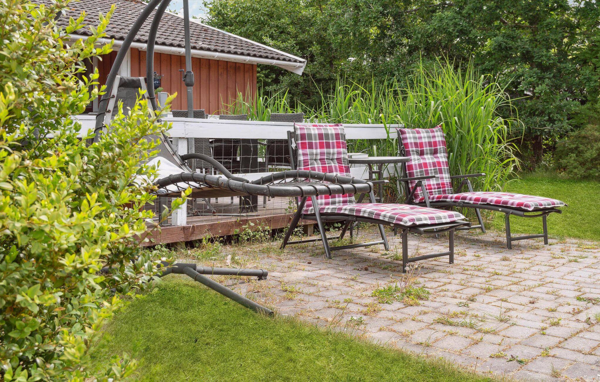 An outdoor seating area with two chairs and a table, surrounded by greenery and a red building in the background.