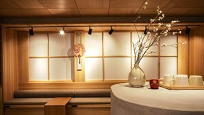 Ein traditioneller japanischer Raum mit Tatami-Böden, einer niedrigen Holzbank, einem runden Tisch mit Blumenvase und einer Schale mit weißen Tassen.
