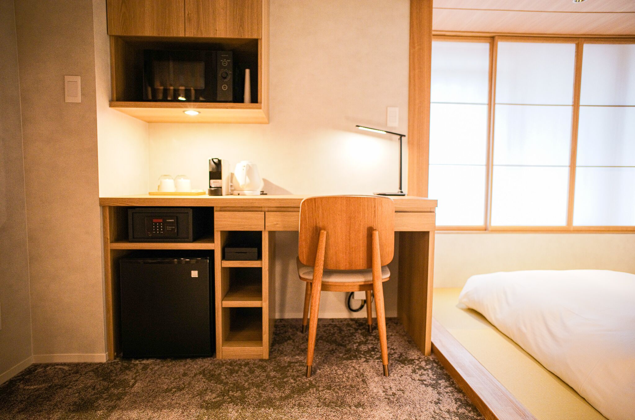 A compact hotel room with a wooden desk, a chair, a bed, and a microwave above the desk.