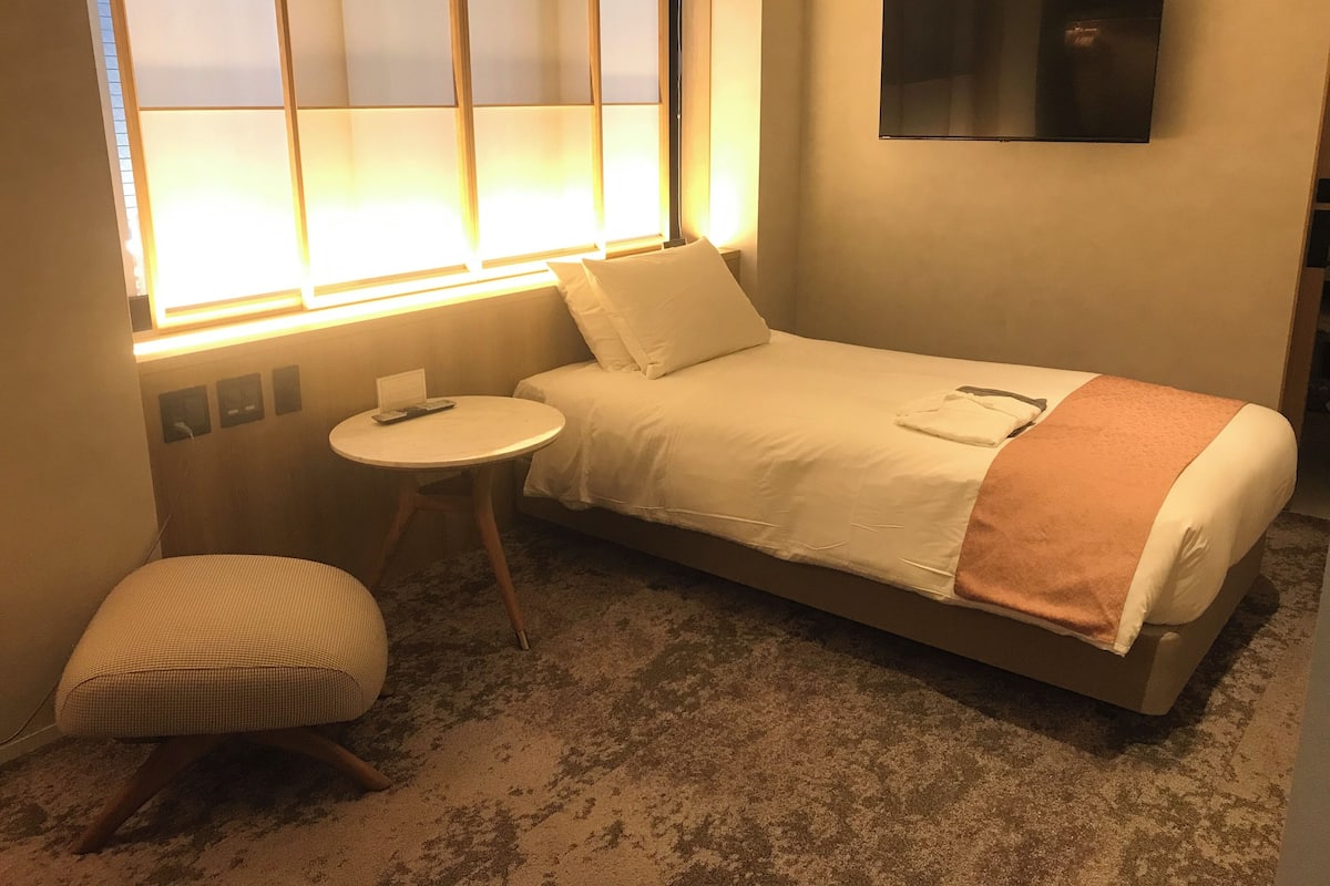 Een enkel bed met een wit-oranje deken, een klein rond tafeltje met een afstandsbediening, een fauteuil en een tv aan de muur.