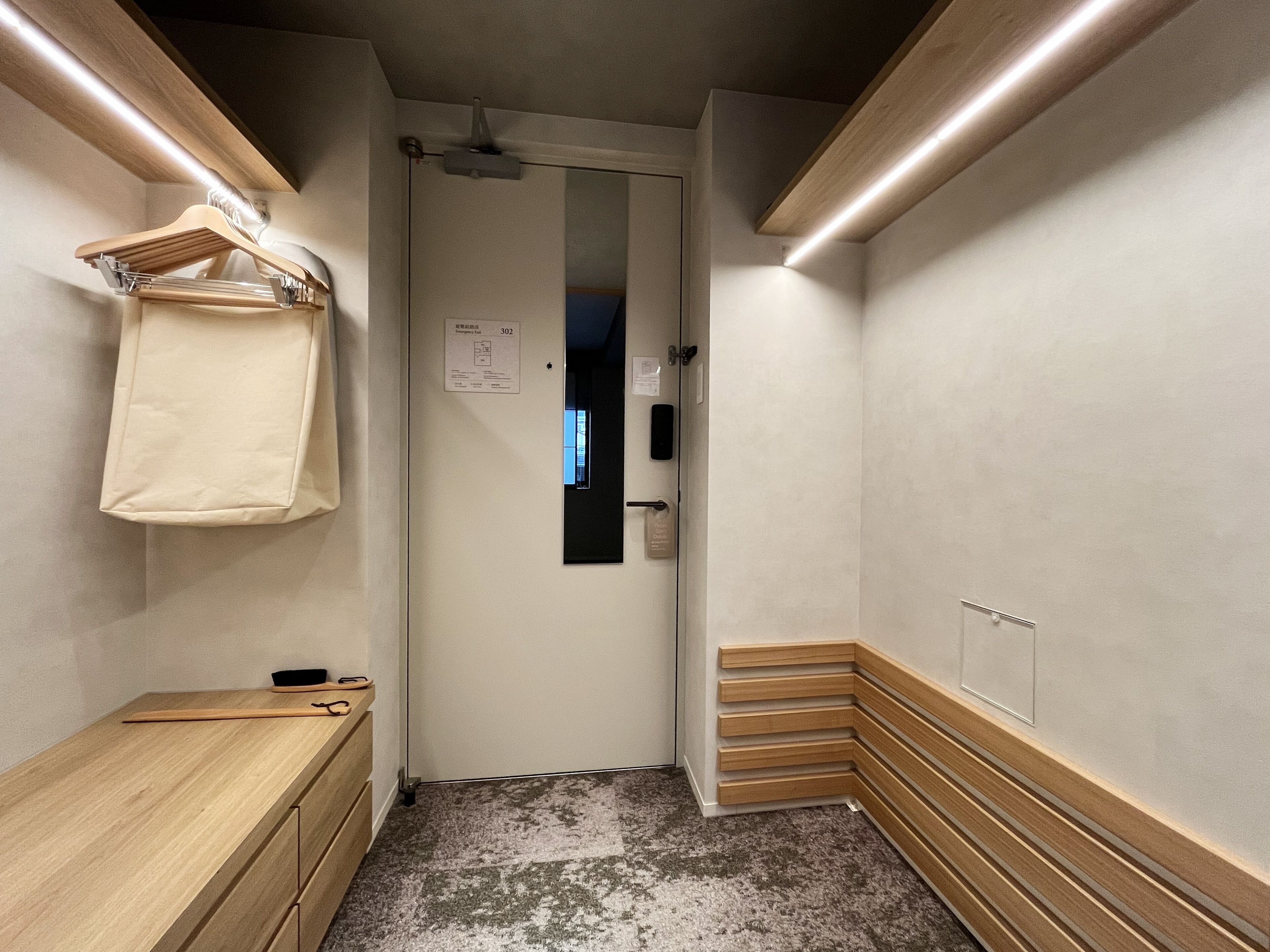 A narrow hallway with a wooden bench on the left, a coat rack with a hanging garment, and a door with a sign on the right.