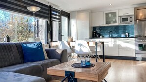 Uma sala de estar moderna com sofá e mesa de centro, uma cozinha com armários brancos e eletrodomésticos de aço inoxidável, e grandes janelas com vista para árvores.