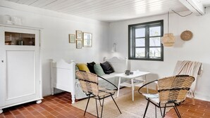 Een knus ingerichte kamer met een wit bed, een tafeltje, rieten stoelen en een raam met uitzicht.