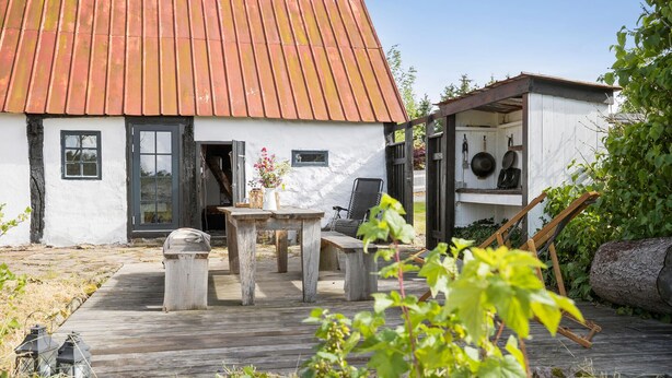 Een rustiek huis met een houten terras, een tafel met stoelen en een kleine tuin.