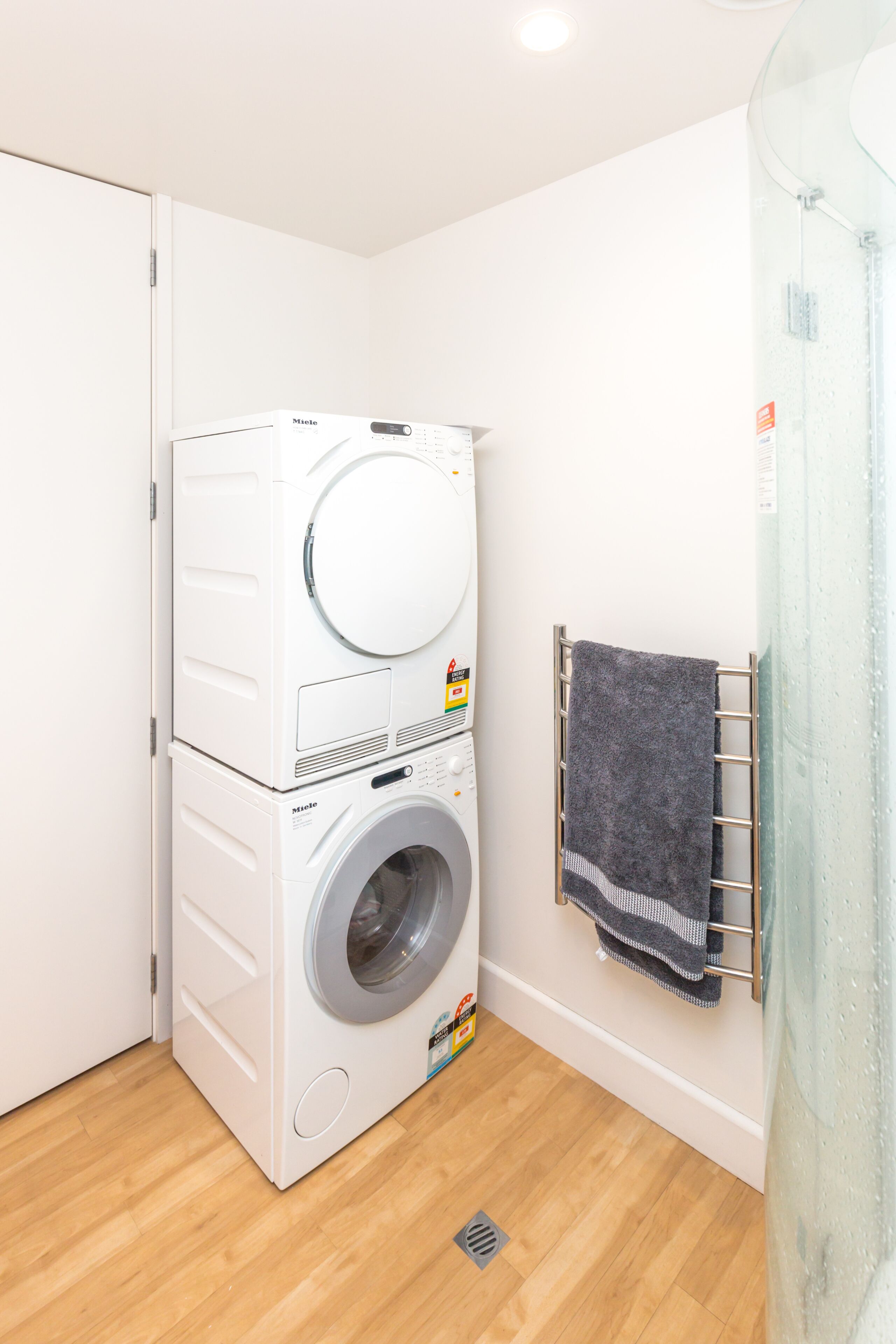 A compact laundry room with a stacked washer and dryer, a metal towel rack, and a wooden floor.