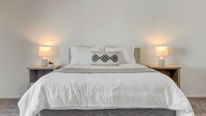 Een netjes opgemaakt bed met witte lakens en een decoratief kussen, met aan weerszijden nachtkastjes met lampen en kleine potplantjes.