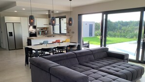 Un salon moderne doté d’un grand canapé d’angle gris, d’un coin repas avec une table blanche et des chaises noires, et d’une cuisine équipée d’appareils électroménagers en acier inoxydable.