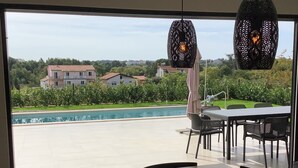 Vue depuis l’intérieur d’une pièce dotée d’une grande fenêtre donnant sur une piscine et un jardin. On y aperçoit une table avec des chaises et un luminaire suspendu.