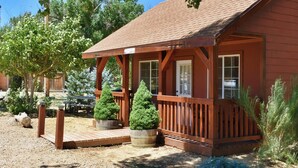 A rustic cabin with a wooden deck, two potted evergreen trees, and a gravel driveway.
