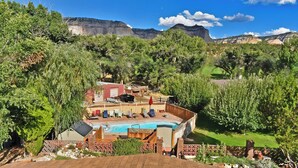 A pool area with lounge chairs and a red umbrella, surrounded by lush greenery and a view of mountains.