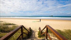 A beach with wooden stairs leading down to the sand, a person with a surfboard, and a clear sky. - Lil Sass on Sassifras - stylish one bedroom apartment, walk to beach (Mudjimba)