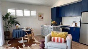 A modern kitchen with blue cabinets, a dining area with a table and chairs, and a living room with a sofa and ottoman. - Lil Sass on Sassifras - stylish one bedroom apartment, walk to beach (Mudjimba)