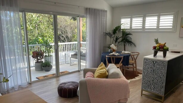 A spacious living room with a sliding glass door leading to a balcony, a wooden dining table, a sofa with cushions, and a decorative cabinet. - Lil Sass on Sassifras - stylish one bedroom apartment, walk to beach (Mudjimba)