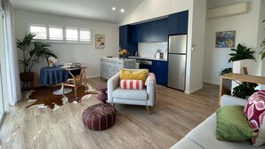 A modern living room with a sofa, a dining table, and a kitchen area with blue cabinets and a refrigerator. - Lil Sass on Sassifras - stylish one bedroom apartment, walk to beach (Mudjimba)