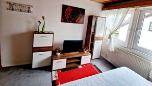 Une chambre avec un plafond en bois, un lit recouvert d’un tapis rouge et blanc, un téléviseur posé sur un meuble en bois, une armoire et une fenêtre avec des rideaux.