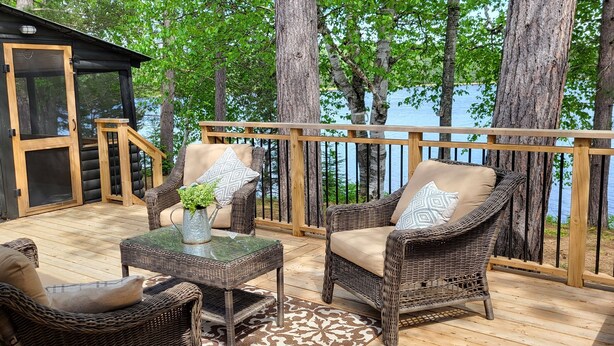 Une cabane en rondins dotée d’une terrasse aménagée avec des meubles en osier, une petite table ornée d’une plante et offrant une vue sur un lac.