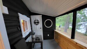 Un petit porche fermé, avec une seule chaise noire et une table, une porte blanche dotée d’une fenêtre circulaire, et une vue sur une zone boisée par le biais d’une grande fenêtre.