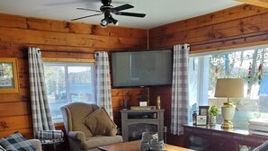 Un salon chaleureux avec des murs en bois, un ventilateur de plafond, un téléviseur à écran plat et une cheminée. On y trouve deux canapés et une table basse.