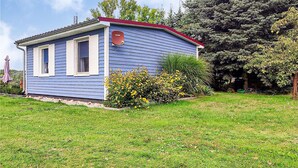 A small, single-story house with a red roof and blue siding, surrounded by a well-maintained lawn and flowering bushes. - Lake front home in Heidesee with WiFi (Heidesee)