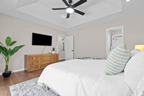 A bedroom with a bed, a ceiling fan, a television on the wall, a wooden dresser, and a potted plant.