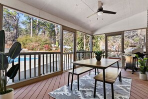 Un patio techado con mesa de madera, bancos, una parrilla y vistas a árboles y una piscina.